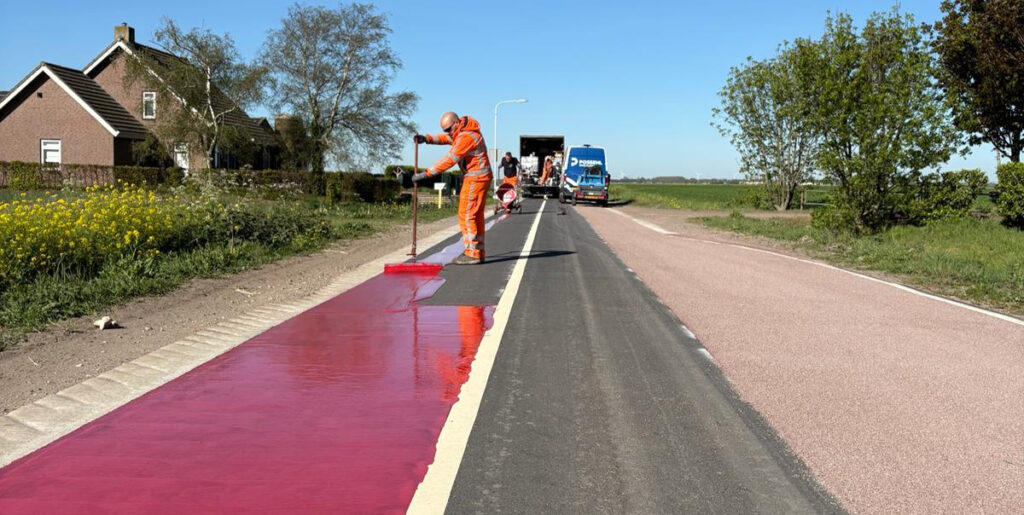Wegwerkers brengen een rode coating aan op een fietspad langs een landelijke weg. Op de achtergrond staat een machine op het asfalt, met bomen en huizen langs de weg onder een heldere blauwe lucht.